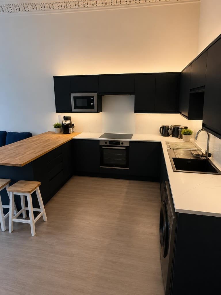 Modern matte black handleless kitchen with white countertops, wooden breakfast bar, and integrated appliances after communal cleaning